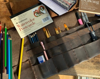 May include: A brown leather roll-up case with multiple pockets for art supplies. The case is open and filled with coloured pencils, paintbrushes, and a watercolour pad. The text on the pad reads "Strathmore Watercolor Postcards".