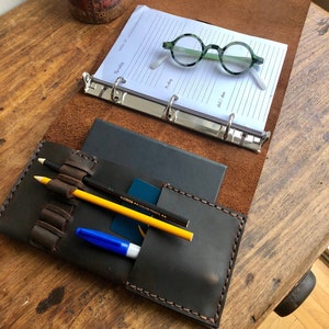 May include: A brown leather binder with a black leather pocket on the front. The pocket has a pen, pencil, and a blue marker inside. The binder has a silver ring mechanism and is open to a blank page with a pair of green glasses on top.