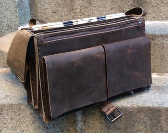 May include: A brown leather messenger bag with a flap closure and multiple pockets. The bag is sitting on a set of concrete stairs.