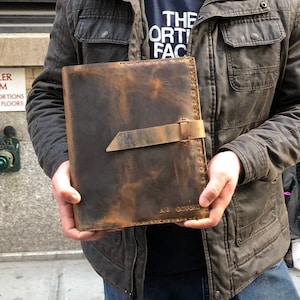 May include: A person wearing a brown jacket and a brown baseball cap is holding a brown leather journal with a strap and the text "AJ GOMONT" embossed on the front.