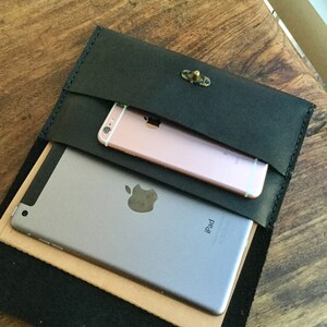 May include: Black leather clutch with a brass clasp, holding a silver iPad and a rose gold smartphone. The clutch is open, revealing the contents. The items are on a wooden surface.