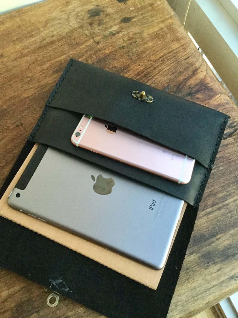 May include: Black leather clutch with a brass clasp, holding a silver iPad and a rose gold smartphone. The clutch is open, revealing the contents. The items are on a wooden surface.