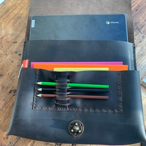 May include: A black leather laptop sleeve with a flap closure and a metal button. The sleeve has a pocket for pencils and a small pocket for papers. The sleeve is open and a laptop with the word "chrome" on the screen is inside.