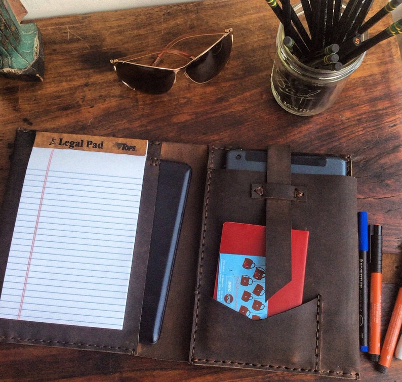 Puede incluir: Un portafolio de cuero marr&oacute;n con un bloc de notas legal, una funda para tableta y un bolsillo para una libreta peque&ntilde;a. El portafolio tiene costuras alrededor de los bordes y una solapa con una correa.