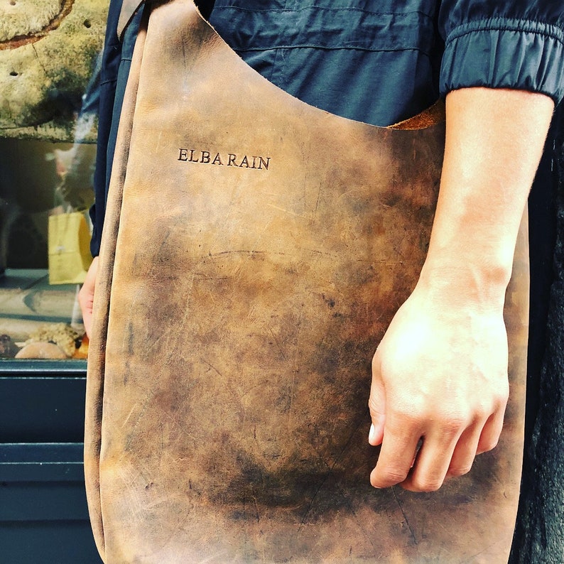 Op de afbeelding: Een bruine leren tote bag met de tekst "ELBA RAIN" erop gegraveerd.