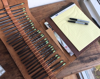 May include: A brown leather pencil roll with 25 black pencils with green tips. The roll is open and a brown leather notepad with a yellow lined notepad is visible. Two black pens are on top of the notepad.