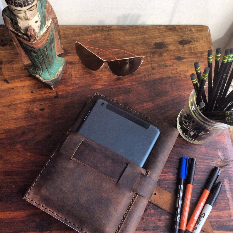 Puede incluir: Un diario de cuero marr&oacute;n con cierre de solapa y un bolsillo para una tableta o tel&eacute;fono. El diario est&aacute; sobre una mesa de madera con una variedad de otros art&iacute;culos, incluyendo un par de gafas de sol, una peque&ntilde;a estatua de madera y un frasco de l&aacute;pices.