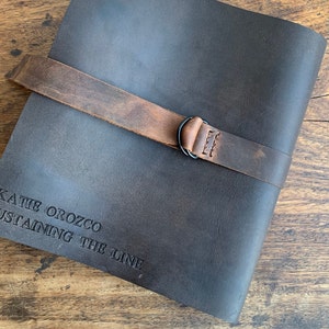 Op de afbeelding: Een donkerbruin lederen dagboek met een bruine leren band en een metalen gesp. Het dagboek heeft de tekst "KATIE OROZCO SUSTAINING THE LINE" gegraveerd op de voorkant.