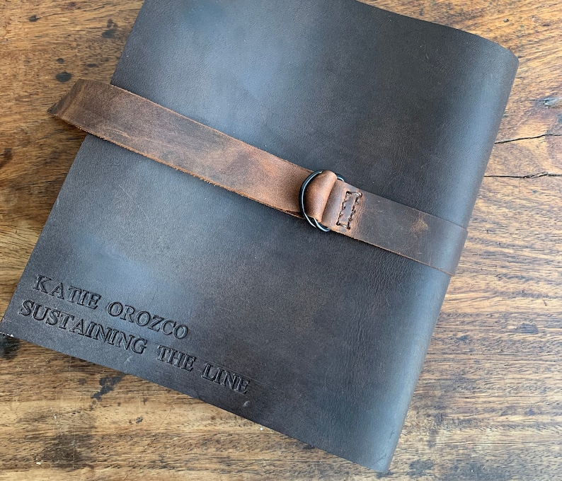 Op de afbeelding: Een donkerbruin lederen dagboek met een bruine leren band en een metalen gesp. Het dagboek heeft de tekst "KATIE OROZCO SUSTAINING THE LINE" gegraveerd op de voorkant.
