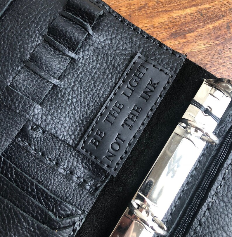 Op de afbeelding: Een zwarte leren planner met een zilveren ringband en een zwart leren label met de tekst "Wees het licht, niet de inkt".