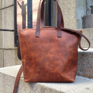 May include: A brown leather tote bag with two top handles and a long strap. The bag has a distressed look and is made of high-quality leather.