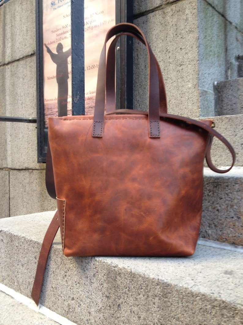 May include: A brown leather tote bag with two top handles and a long strap. The bag has a distressed look and is made of high-quality leather.