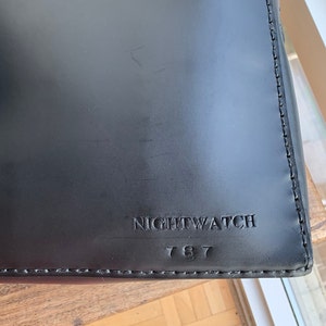 Op de afbeelding: Zwarte leren tas met een gestikte rand en de tekst "NIGHTWATCH 787" gegraveerd op de voorkant.