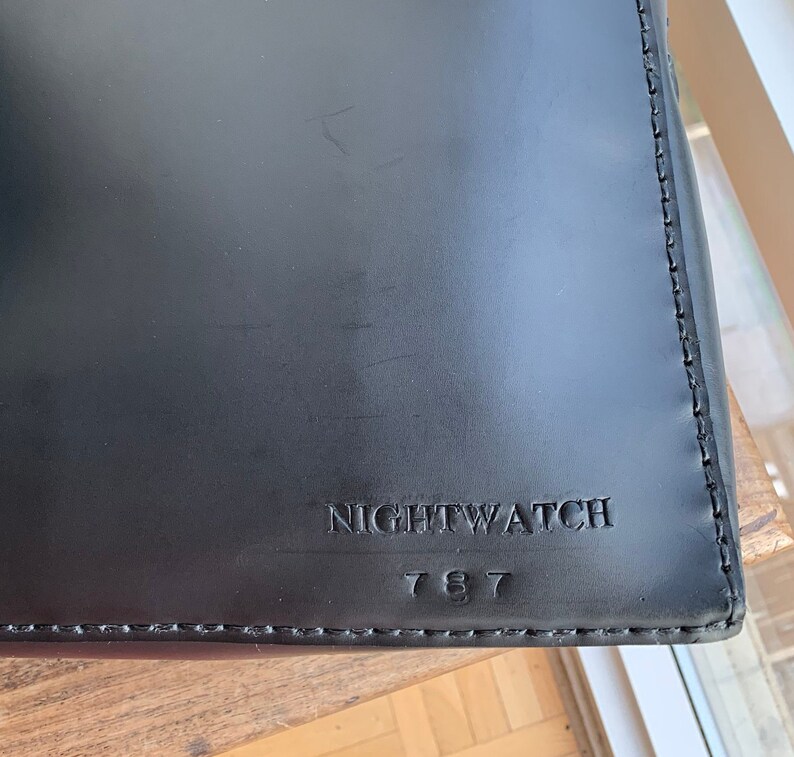 Op de afbeelding: Zwarte leren tas met een gestikte rand en de tekst "NIGHTWATCH 787" gegraveerd op de voorkant.