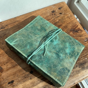 May include: A teal leather journal with a light green leather tie closure. The journal has a distressed look and is lying on a wooden surface.