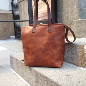 May include: A brown leather tote bag with a long strap. The bag has a stitched design and a worn look.