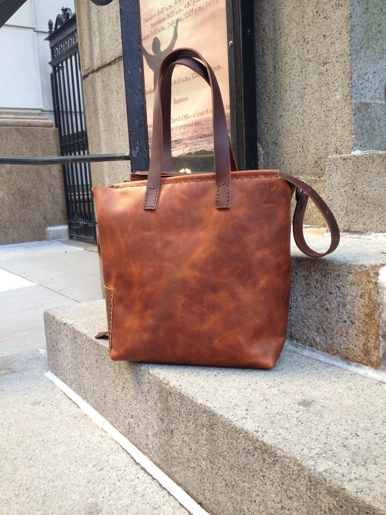 May include: A brown leather tote bag with a long strap. The bag has a stitched design and a worn look.