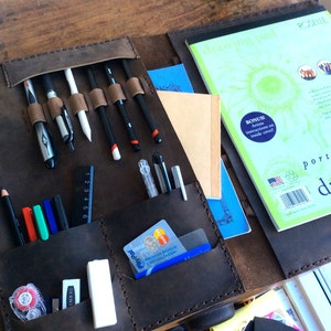 May include: A brown leather portfolio case with multiple pockets and slots for holding art supplies, including pens, pencils, markers, and a ruler. The case also has a pocket for a credit card. The case is open and the inside is visible. The case is labelled "drawing pad" and "portfolio series".