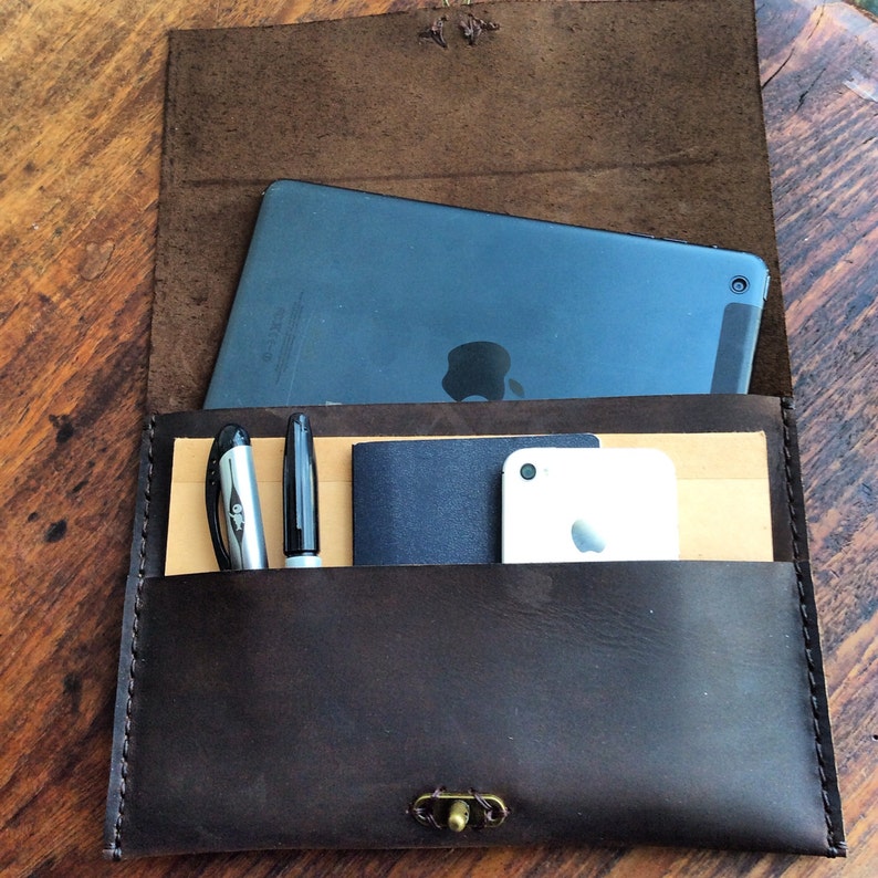 May include: A dark brown leather folio with a flap, holding a tablet, pens, a small notebook, and a smartphone. The folio has a brass clasp and is stitched along the edges. The interior has a light brown pocket.
