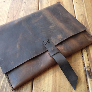 May include: A brown leather laptop sleeve with a dark brown strap and a stitched closure. The sleeve has a distressed look and is made of high-quality leather.