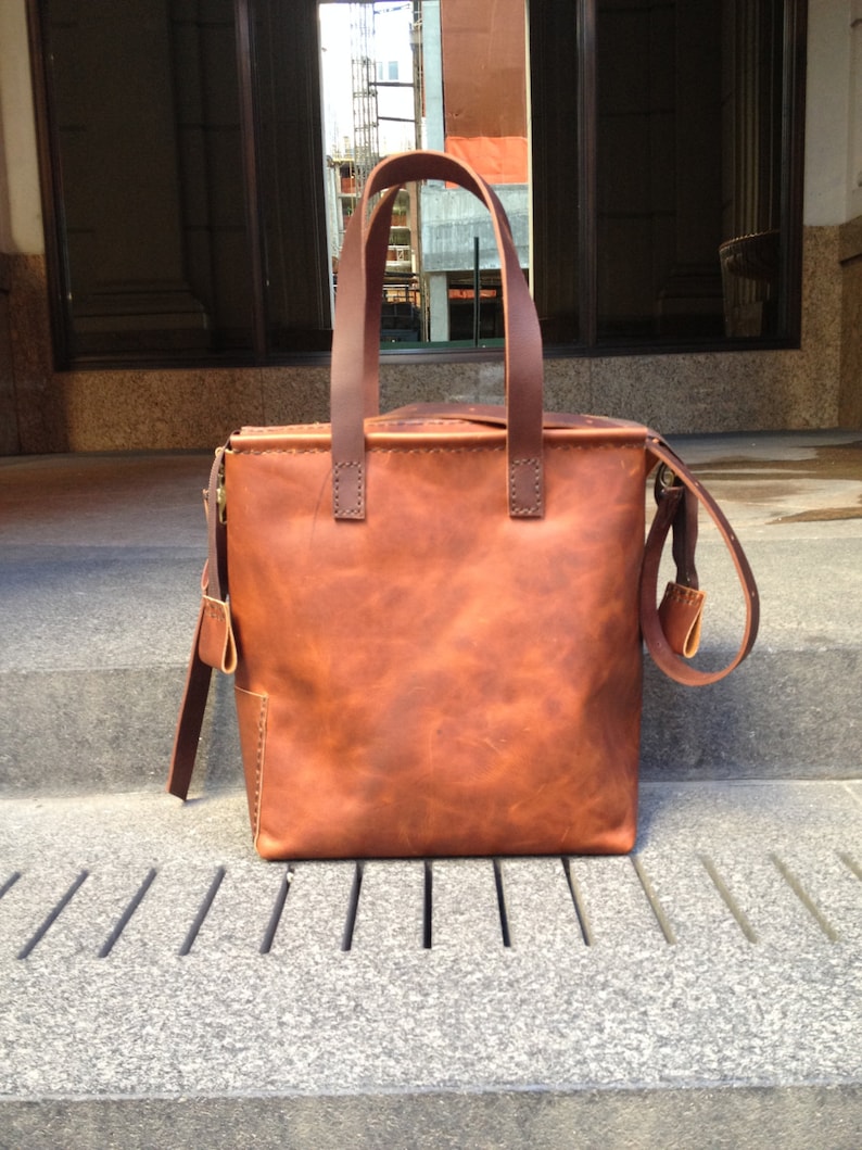 May include: A brown leather tote bag with a top zipper closure and a long strap. The bag has a distressed look and is made of high-quality leather.