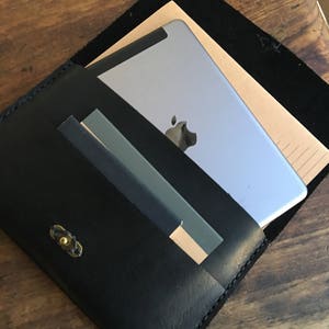May include: Black leather folio case with a brass clasp, holding a silver tablet and a notepad. The case is open, revealing the contents on a wooden surface. The folio has a minimalist design.