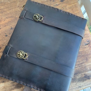 May include: A dark brown leather journal with two straps and brass clasps. The journal is closed and lying on a wooden surface.