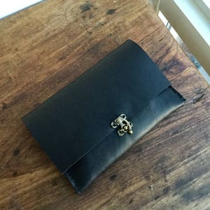 May include: A black leather clutch with a brass clasp. The rectangular clutch is closed and lies on a wooden surface. The clasp is ornate and adds a vintage touch to the accessory.