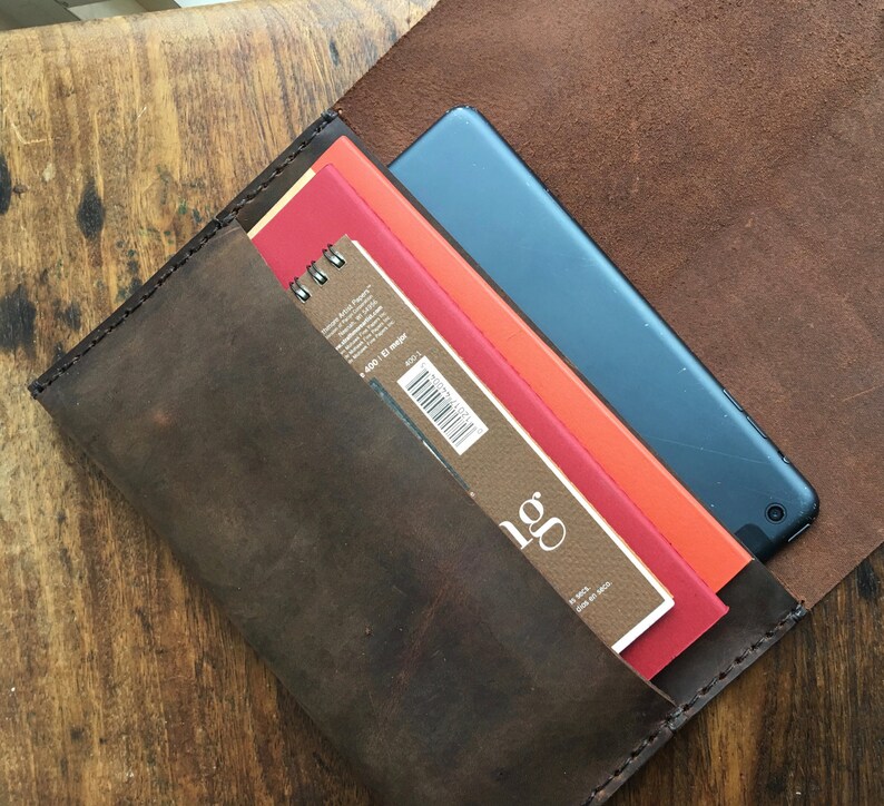 May include: A dark brown leather case, open to reveal a blue tablet, and several art pads in red, orange, and brown. The case is made of distressed leather and has visible stitching.