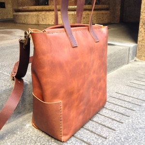 May include: A brown leather tote bag with a long strap and a smaller pocket on the front. The bag has a worn, distressed look.