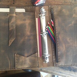 May include: A brown leather binder with a zippered pocket, a flap for a tablet, and a ring binder with colored pencils inside.