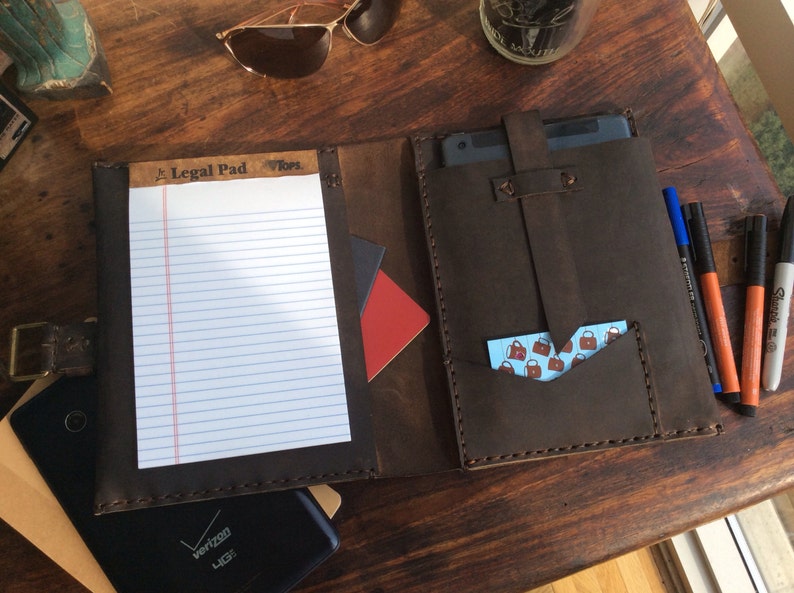 Puede incluir: Un portafolio de cuero marr&oacute;n con un bloc de notas, un bolsillo para una tableta y un bolsillo m&aacute;s peque&ntilde;o para tarjetas de visita. El portafolio tiene un borde cosido y una solapa con el texto "Legal Pad" en &eacute;l.