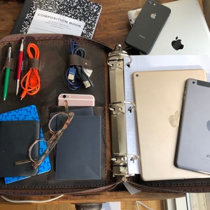 Puede incluir: Un maletín de cuero marrón con cierre de cremallera. El maletín está abierto y contiene una variedad de artículos, incluyendo una carpeta, dos tabletas, un teléfono, bolígrafos y cables. El maletín está sobre una mesa de madera.