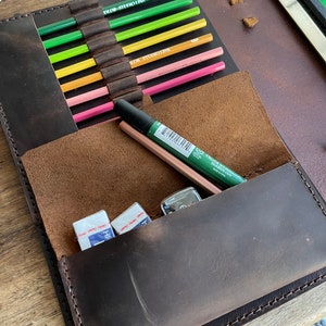 Puede incluir: Un portafolio de cuero marrón con una solapa que se abre para revelar una variedad de artículos de arte, incluidos lápices de colores, marcadores y borradores. El portafolio tiene un borde cosido y un cierre de solapa.