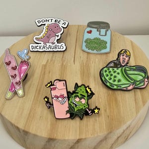 May include: A collection of enamel pins featuring various designs. Included are a pink dinosaur pin with the text "DON'T BE A DICKASAURUS", a jar of green buds, a pink lighter with a smiling face, and a green cannabis character. Also included are two pink joints and a person in a green croc.
