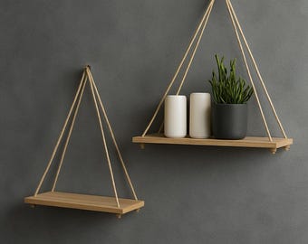 May include: Two wooden hanging shelves with natural rope supports against a gray wall. One shelf holds two white cylindrical candles and a black potted plant. The other shelf is empty. The shelves have a minimalist, modern design.