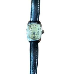 May include: A rectangular-faced wristwatch with a black leather band and white stitching. The watch face is a light cream color with black numerals and hands. The watch has a silver-colored case and a small crown.