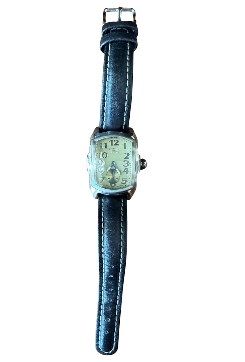 May include: A rectangular-faced wristwatch with a black leather band and white stitching. The watch face is a light cream color with black numerals and hands. The watch has a silver-colored case and a small crown.
