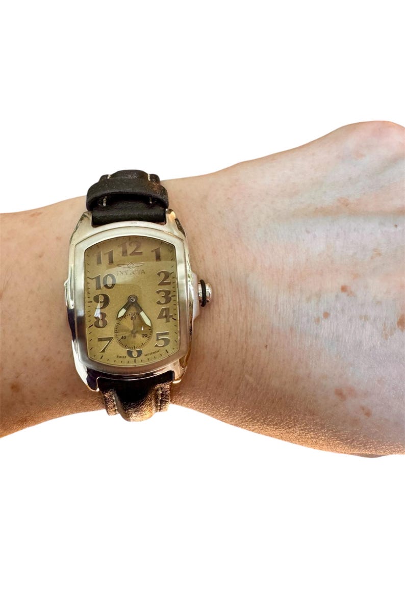 May include: A rectangular Invicta watch with a silver-tone case and a brown leather strap. The watch face is gold with black numerals and hands. The watch is on a wrist.