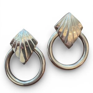 May include: A pair of silver-toned earrings. Each earring features a geometric, fan-shaped top attached to a circular hoop. The earrings are displayed against a white background, highlighting their metallic sheen and design.