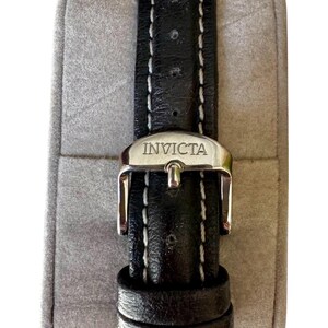 May include: Black leather watch band with white stitching and a silver buckle. The buckle is engraved with the brand name "INVICTA". The watch band is displayed on a gray, padded surface.