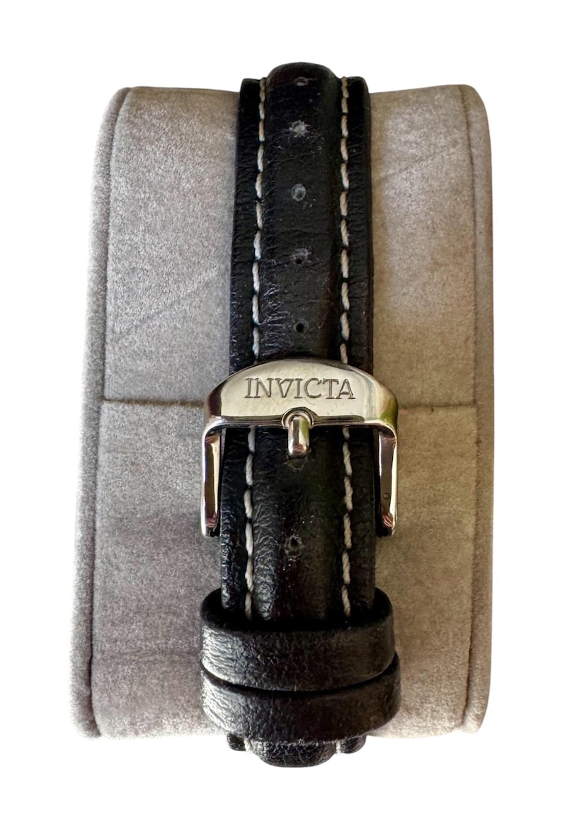 May include: Black leather watch band with white stitching and a silver buckle. The buckle is engraved with the brand name "INVICTA". The watch band is displayed on a gray, padded surface.