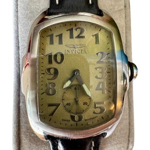May include: A silver-toned Invicta watch with a rectangular face and a black leather band. The watch face is a light olive green with large black numerals and a small subdial. The watch is marked "SWISS MOVEMENT".