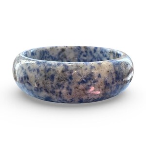 May include: A round ring made of a speckled blue and gray stone. The ring has a smooth, polished surface and a wide band. The stone's pattern features dark blue spots and swirls against a lighter gray background. The ring is a piece of jewelry.