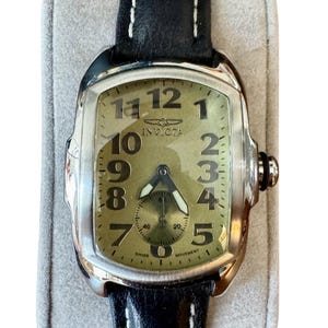 May include: A rectangular Invicta watch with a black leather band and white stitching. The watch face is a light olive green with black numerals and hands. The watch has a small subdial and the word "Invicta" is printed on the face.