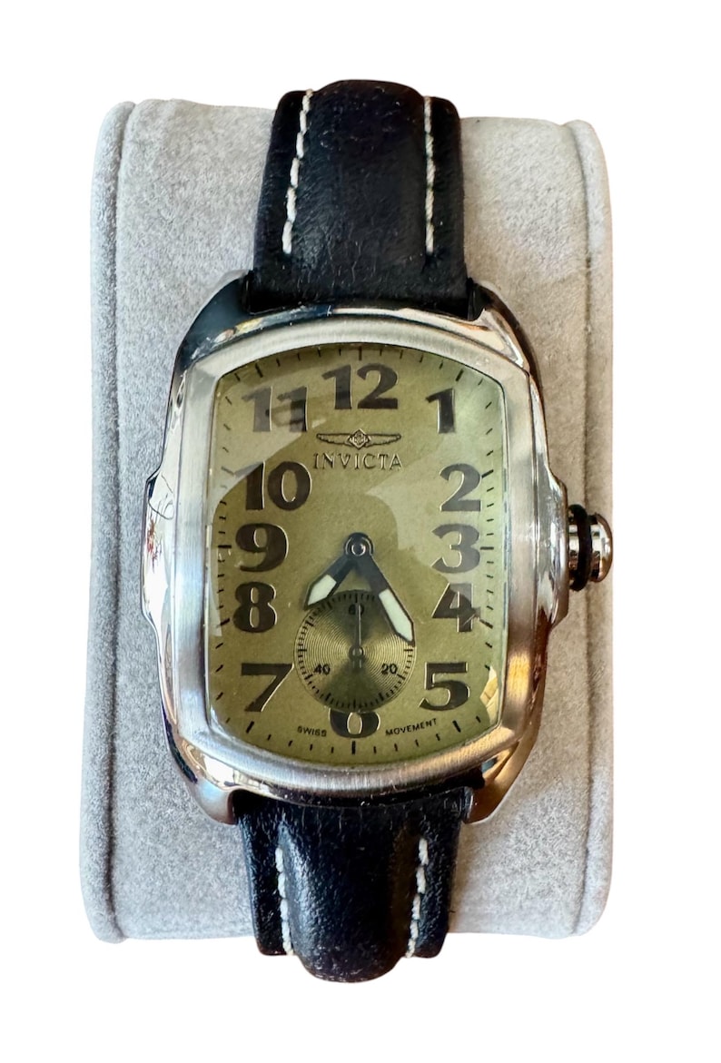 May include: A rectangular Invicta watch with a black leather band and white stitching. The watch face is a light olive green with black numerals and hands. The watch has a small subdial and the word "Invicta" is printed on the face.