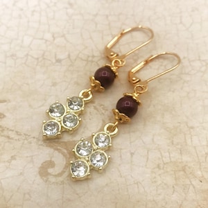 May include: A pair of gold dangle earrings with a cluster of clear rhinestones and a single maroon pearl bead on each earring.