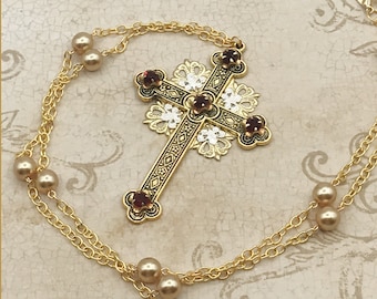 May include: A gold chain necklace with a large ornate cross pendant featuring red gemstones. The chain is adorned with small pearl-like beads.