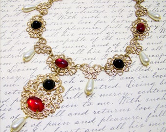 May include: A gold filigree necklace with red and black gemstones and pearl accents. The necklace is on a white background with cursive writing. The text reads "Treasures for a Queen".