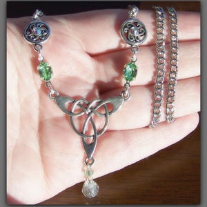 Puede incluir: Un collar con un colgante de nudo celta plateado, acentos de piedras preciosas verdes y una gota de cristal. El collar est&aacute; en una cadena de plata.